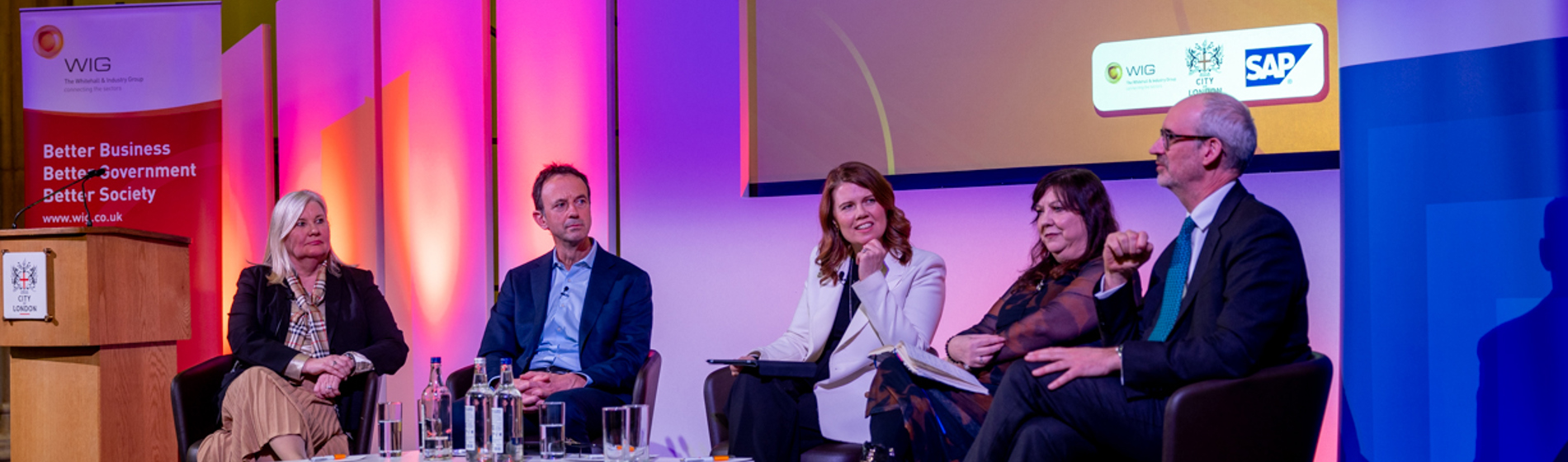 Five speakers participate in a WIG panel discussion, seated on stage with pink and purple lighting.