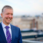Professional man in a suit smiling on a London rooftop, with the Palace of Westminster visible.