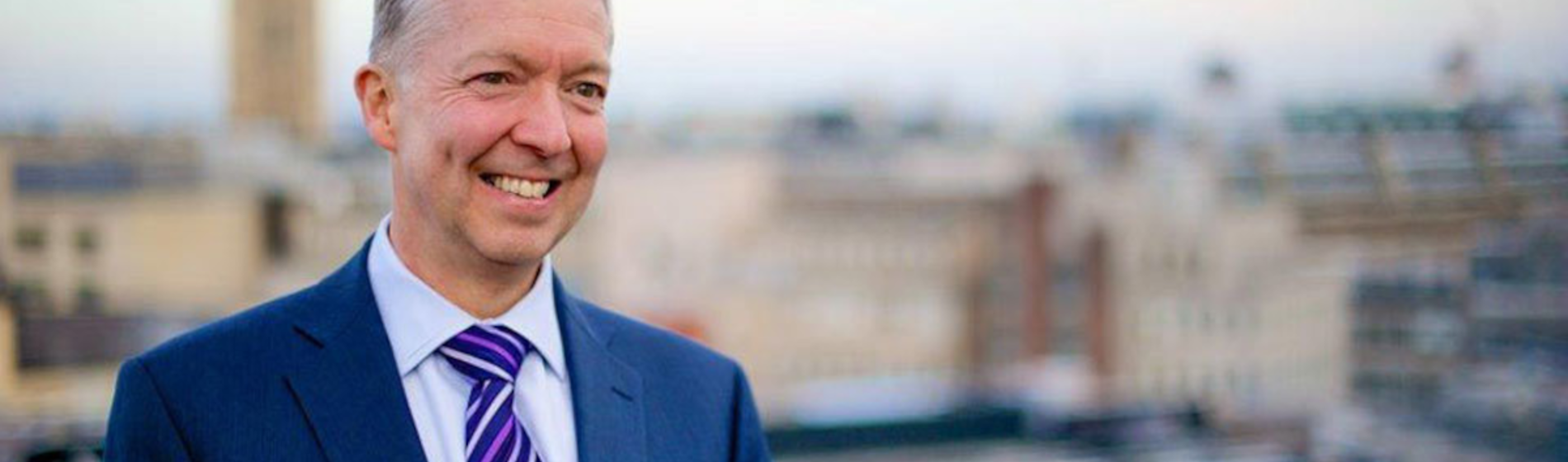 Professional man in a suit smiling on a London rooftop, with the Palace of Westminster visible.