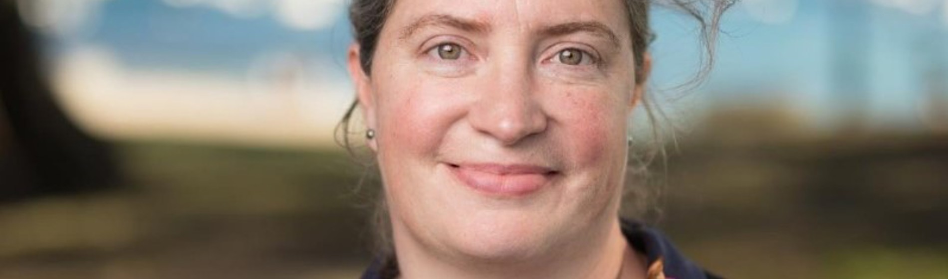 Headshot of a smiling woman with greyish-brown hair, blurred outdoor setting.