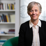 A smiling professional woman with short grey hair, wearing a black jacket, against a bookshelf background.
