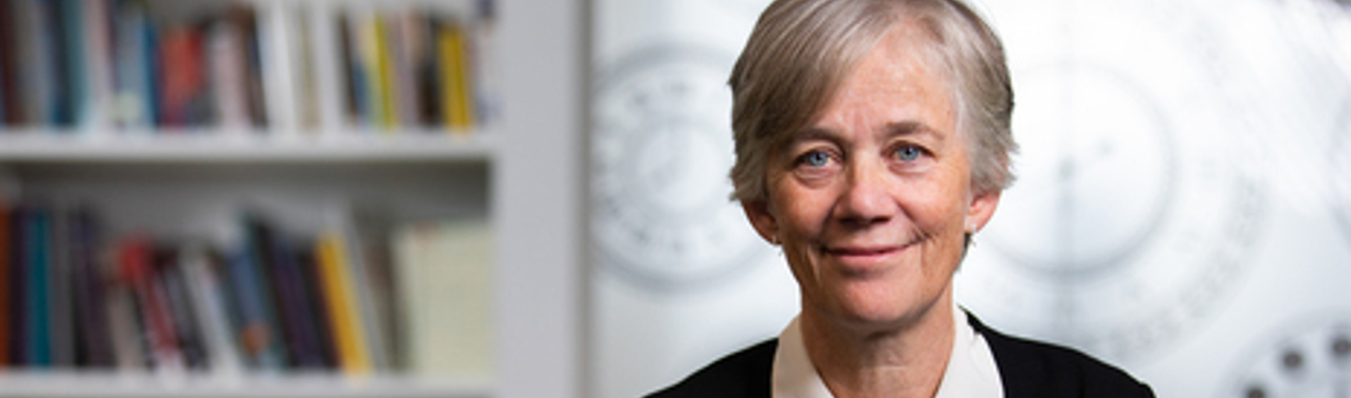 A smiling professional woman with short grey hair, wearing a black jacket, against a bookshelf background.