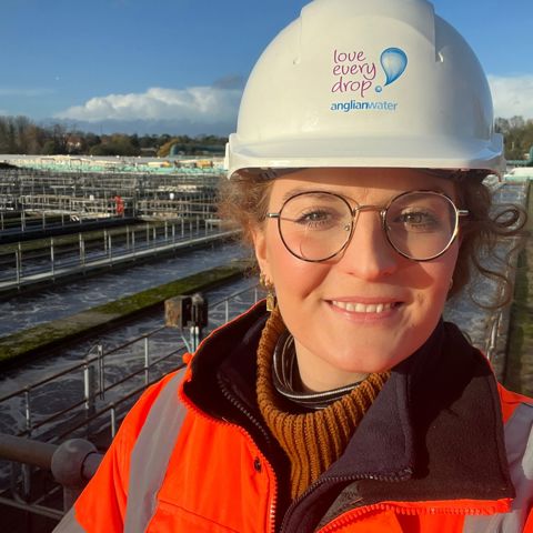 Smiling worker in hi-vis jacket and Anglian Water hard hat at a water treatment facility.