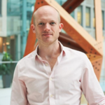 A bald man in a pink shirt stands in a modern office atrium.