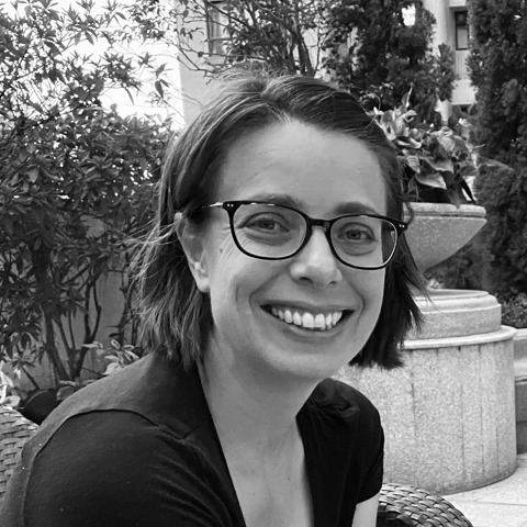 Black and white portrait of a smiling woman in glasses, seated outdoors amongst foliage.