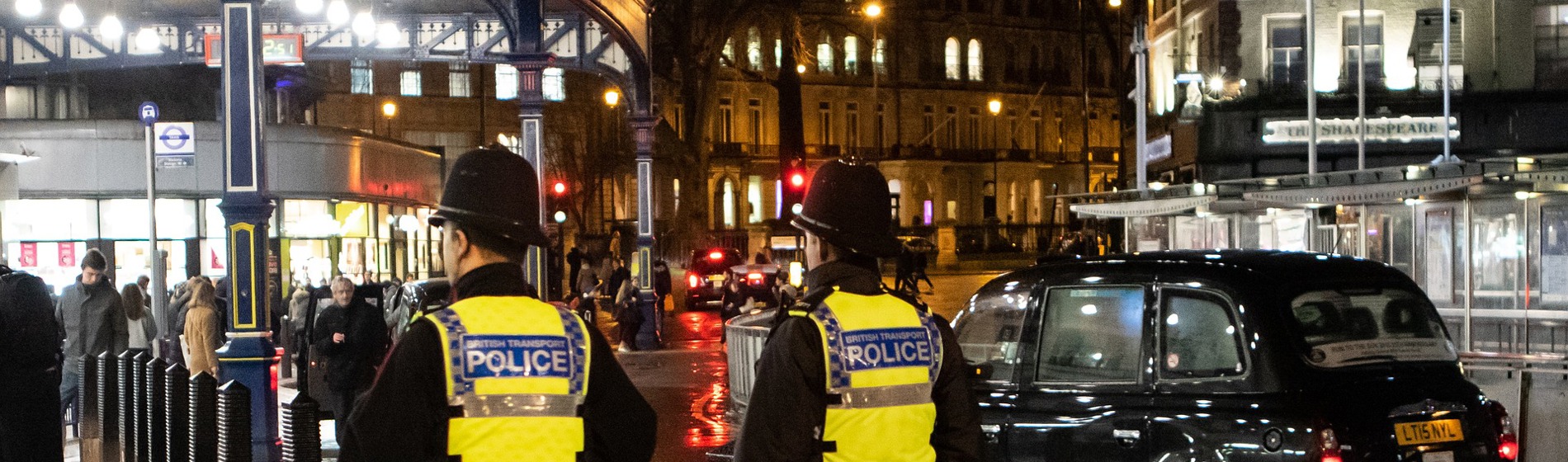 Two police officers in high-vis vests and helmets stand near a black London taxi at night.