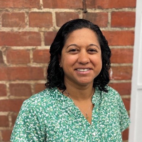 A smiling woman with dark hair wearing a green floral shirt against a brick wall.