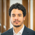 Portrait of a man with dark, curly hair and a beard, wearing a suit jacket.