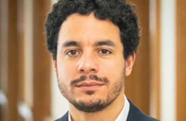 Portrait of a man with dark, curly hair and a beard, wearing a suit jacket.