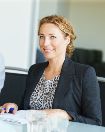 A smiling businesswoman in a dark blazer and leopard print top, with a colleague seated behind her.