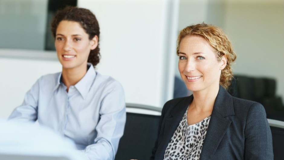 A smiling businesswoman in a dark blazer and leopard print top, with a colleague seated behind her.