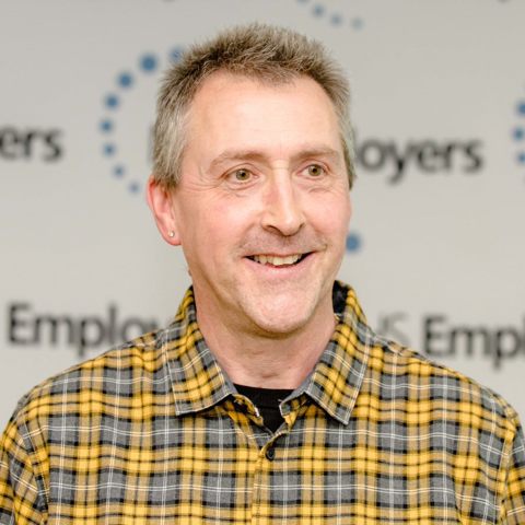 Smiling man in a yellow and grey checked shirt, standing against a branded background.