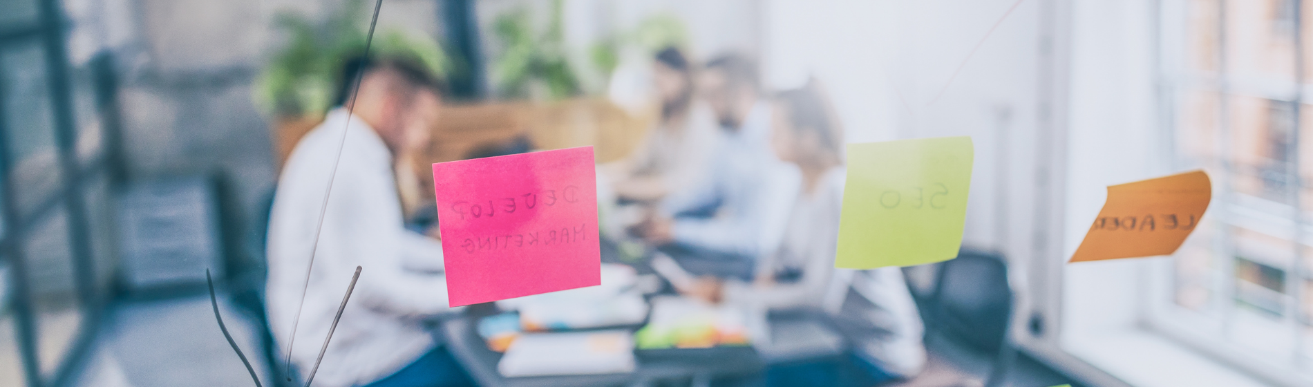 Sticky notes reading "Marketing" and "SEO" on a glass wall, with blurred colleagues meeting in the background.